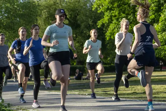 En grupp kvinnor tränar löpning utomhus. De springer i ett parkliknande område, redo att springa 1 km tid.
